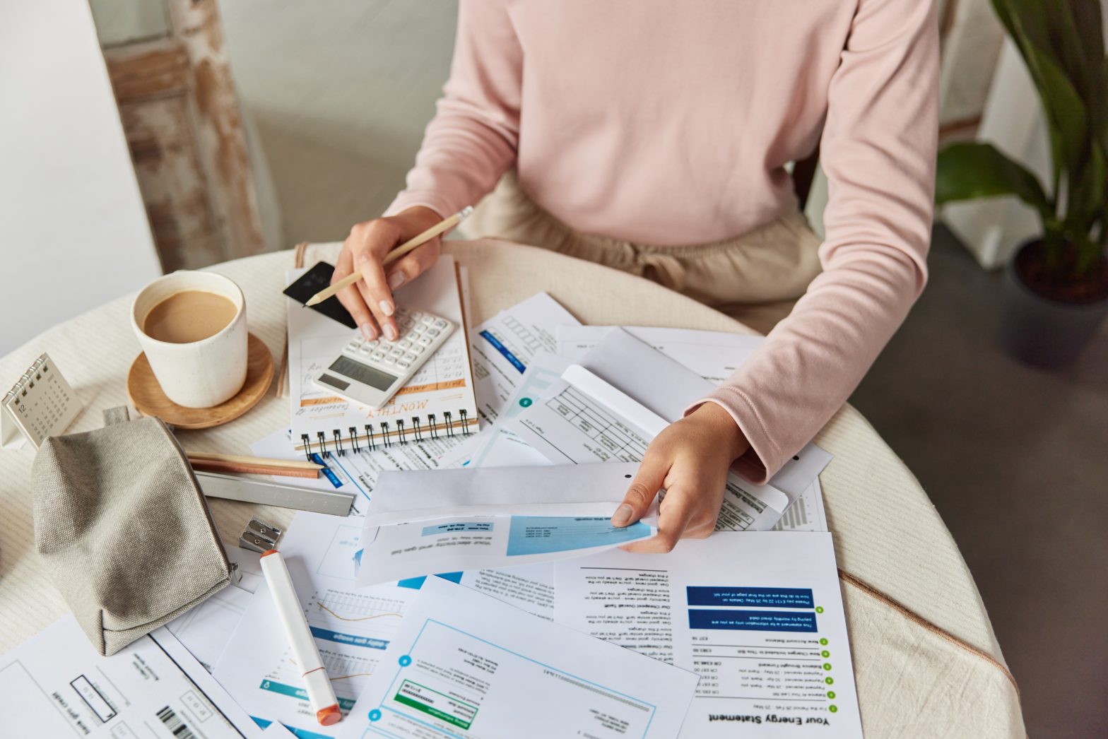 Woman analyses bills while sitting at a round table with paper documents, a calculator and coffee.