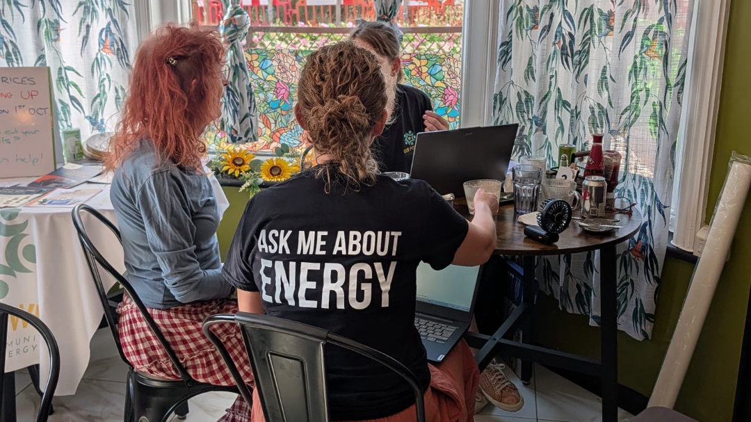 The interior of a cafe with floral curtains. Three women are sitting around a table, talking. One of them had her back to the camera and is wearing a black t-shirt that says "Ask me about energy"