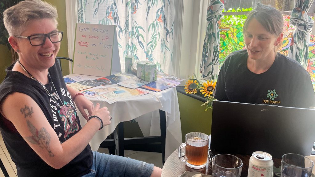 Two women sitting down at a table inside a cafe, smiling, mid-conversation. One of them is wearing a black t-shirt branded with the Our Power logo. Behind them, a hand written sign says "Has prices are going up again on 1st Oct. Want to cut your bills? We can help!"